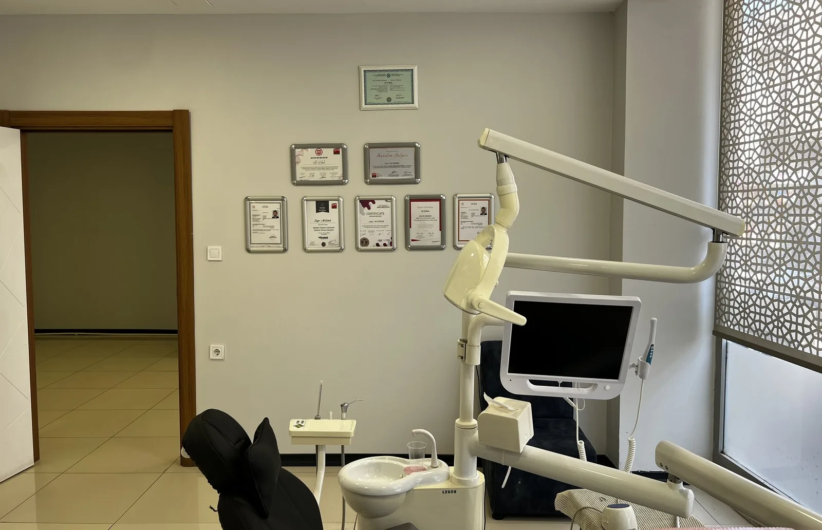 Kliniğimizin tedavi odalarından biri: diş ünitesi, hasta ekranı ve duvarda hekimlerimizin diploma ve katılım belgeleri